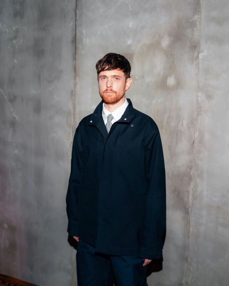 Portrait of a man with short brown hair and a trimmed beard, wearing a dark jacket over a white shirt and tie, standing against a minimalist concrete wall background. Professional and modern aesthetic with muted tones.