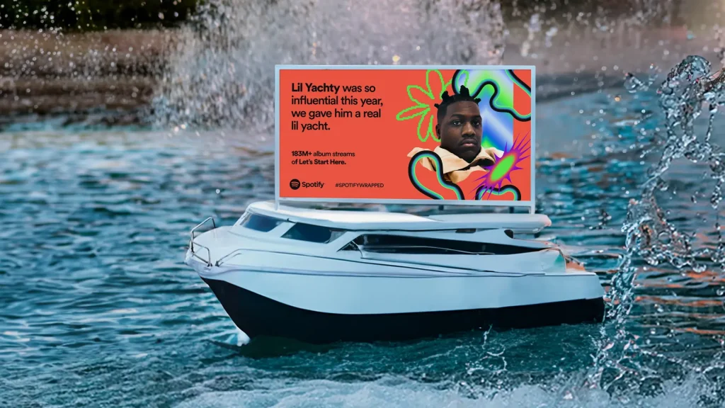 Picture of a billboard on top of a small motorboat in the water. The billboard features a man and a company logo. 